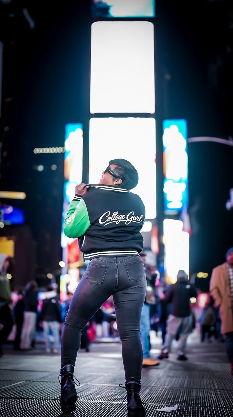 Jessica in Times Square at night wearing a College Gurl varsity jacket and black Sunnies